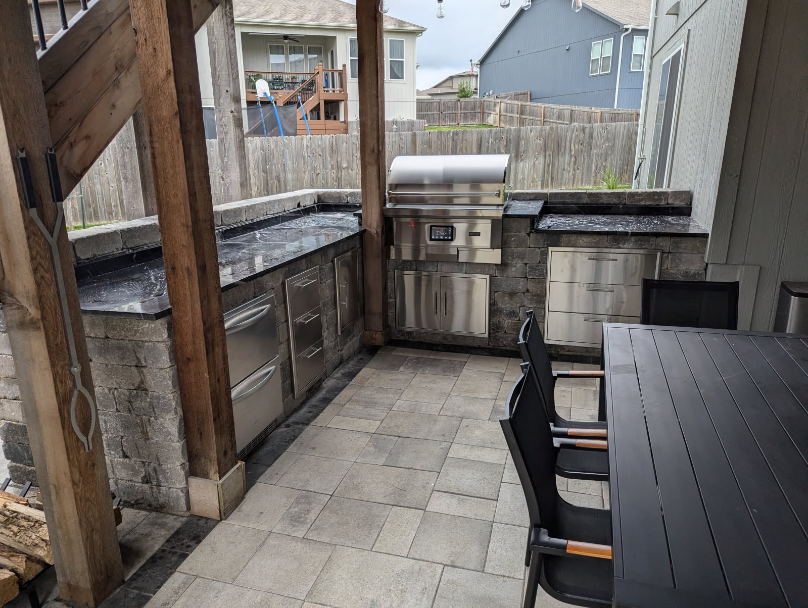 Outdoor kitchen with stainless steel appliances, covered by wooden beams. Patio furniture nearby, surrounded by a wooden fence and residential houses.