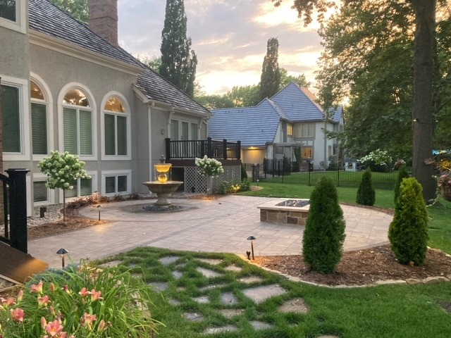 Elegant backyard features a paved patio with a fountain, fire pit, manicured lawn, and flowering plants next to a charming house at sunset.