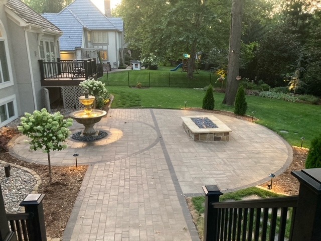 A well-maintained backyard with a paved patio, central fountain, and fire pit, surrounded by trees and greenery, creating a serene outdoor space.