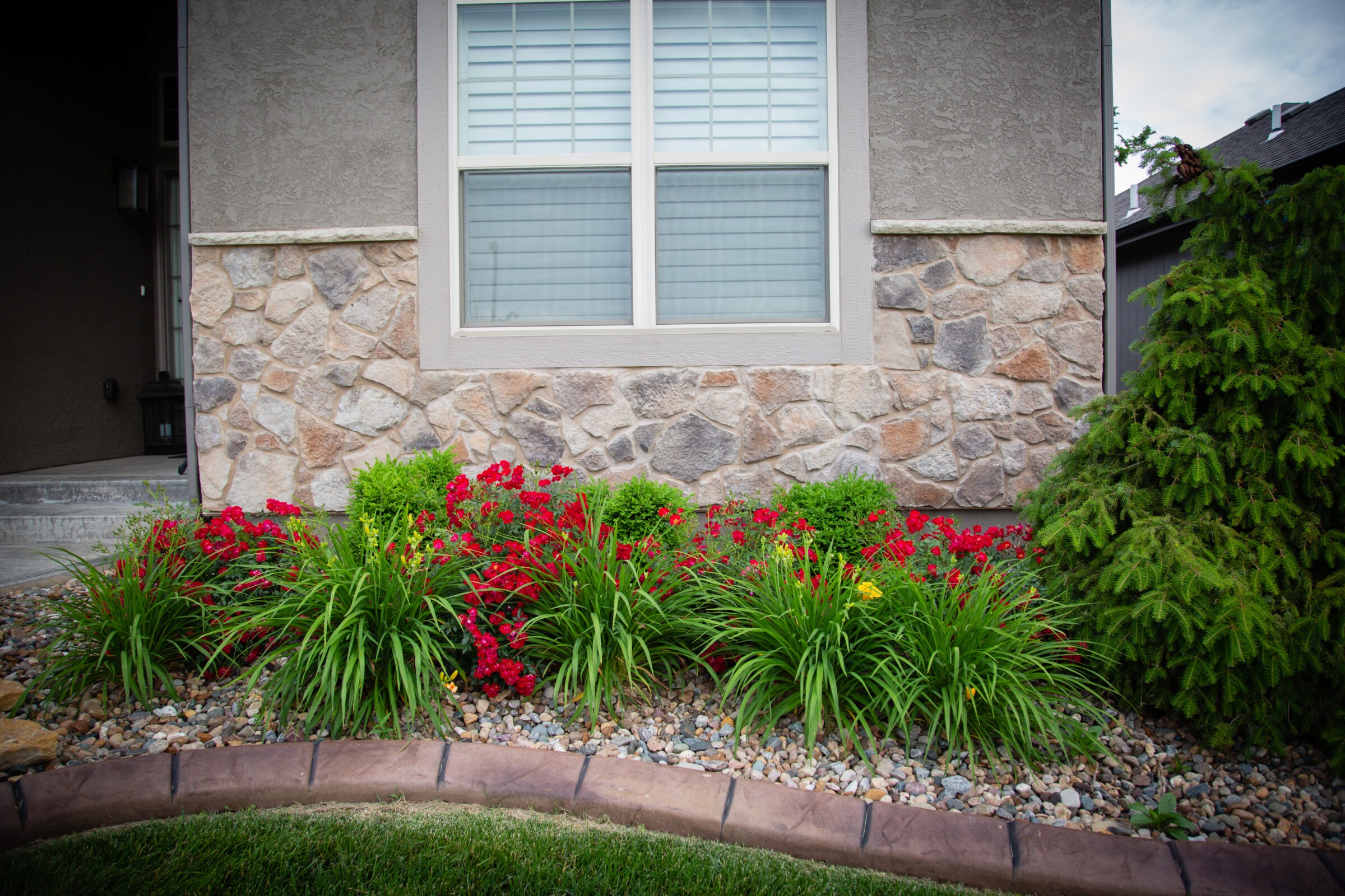 A stone house exterior with a manicured garden featuring red flowers, green shrubs, and a small tree, bordered by decorative stones.