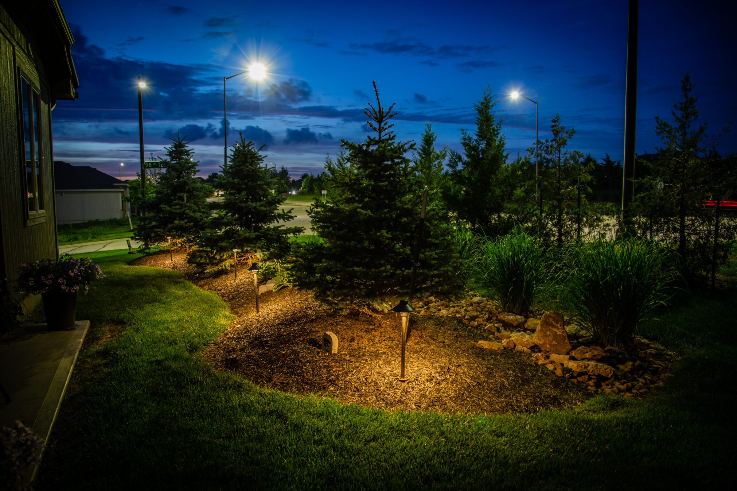 A landscaped garden with illuminated trees and path lights under a twilight sky, surrounded by grass and decorative plants near a building.