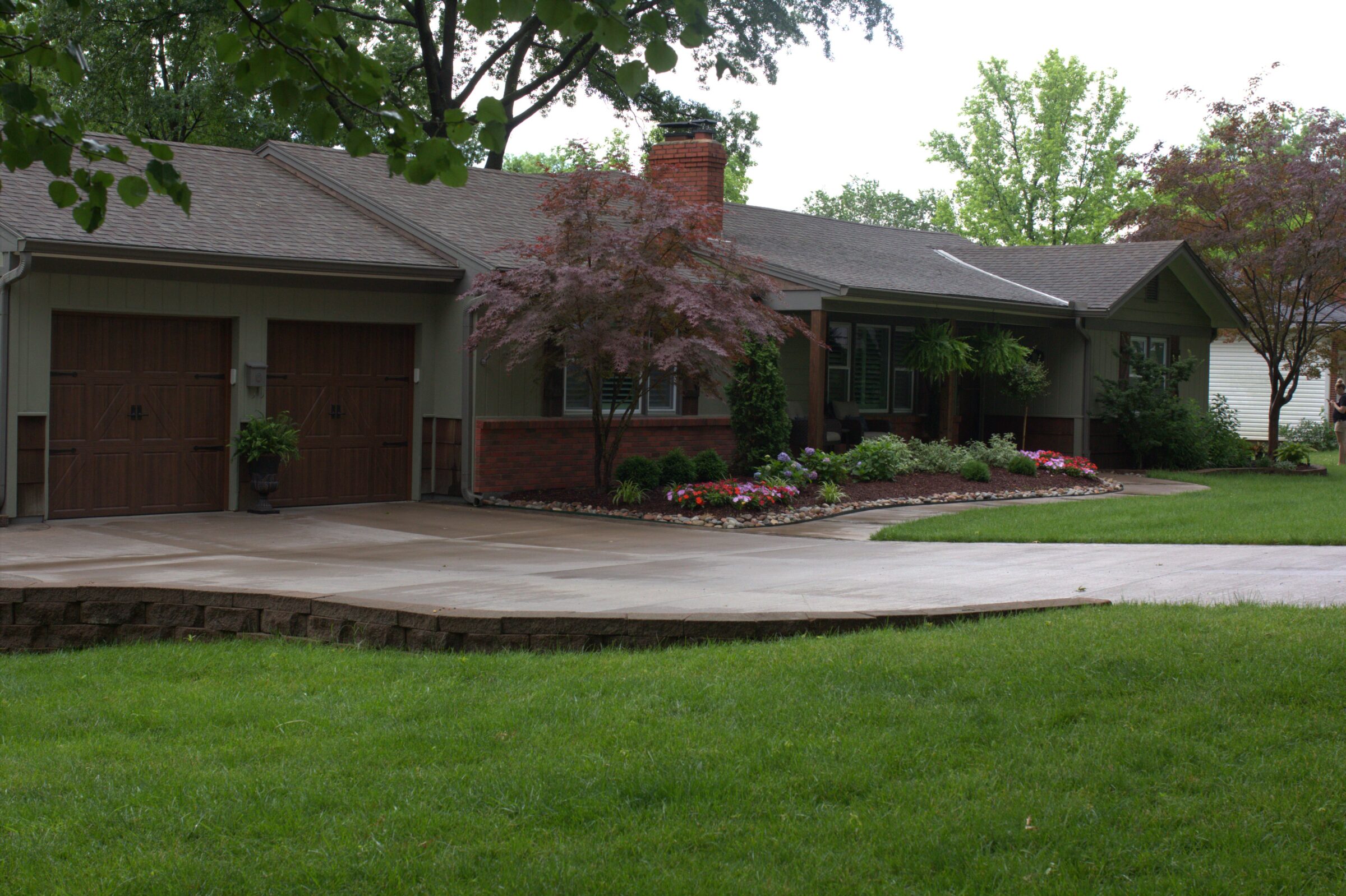 Welcoming front yard with curved concrete driveway, manicured lawn, and vibrant garden beds accented by ornamental trees and foundation plantings.