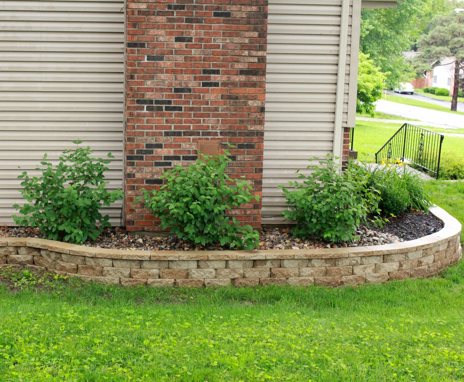 Neatly landscaped garden bed bordered by a curved stone retaining wall, featuring low-maintenance shrubs and decorative river rock for added texture.