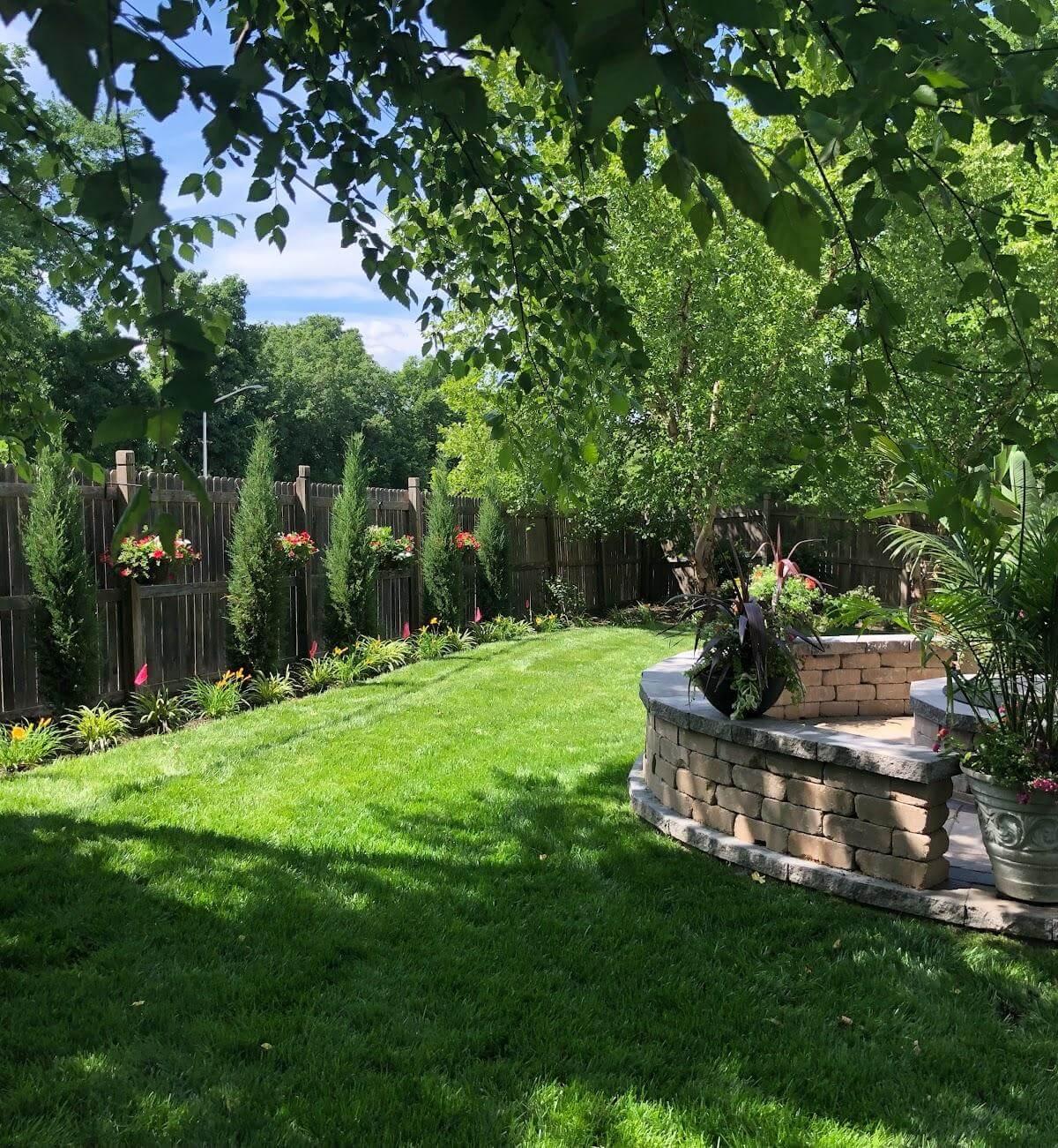 Backyard landscape with raised stone patio, manicured lawn, privacy fence, and colorful flower beds.
