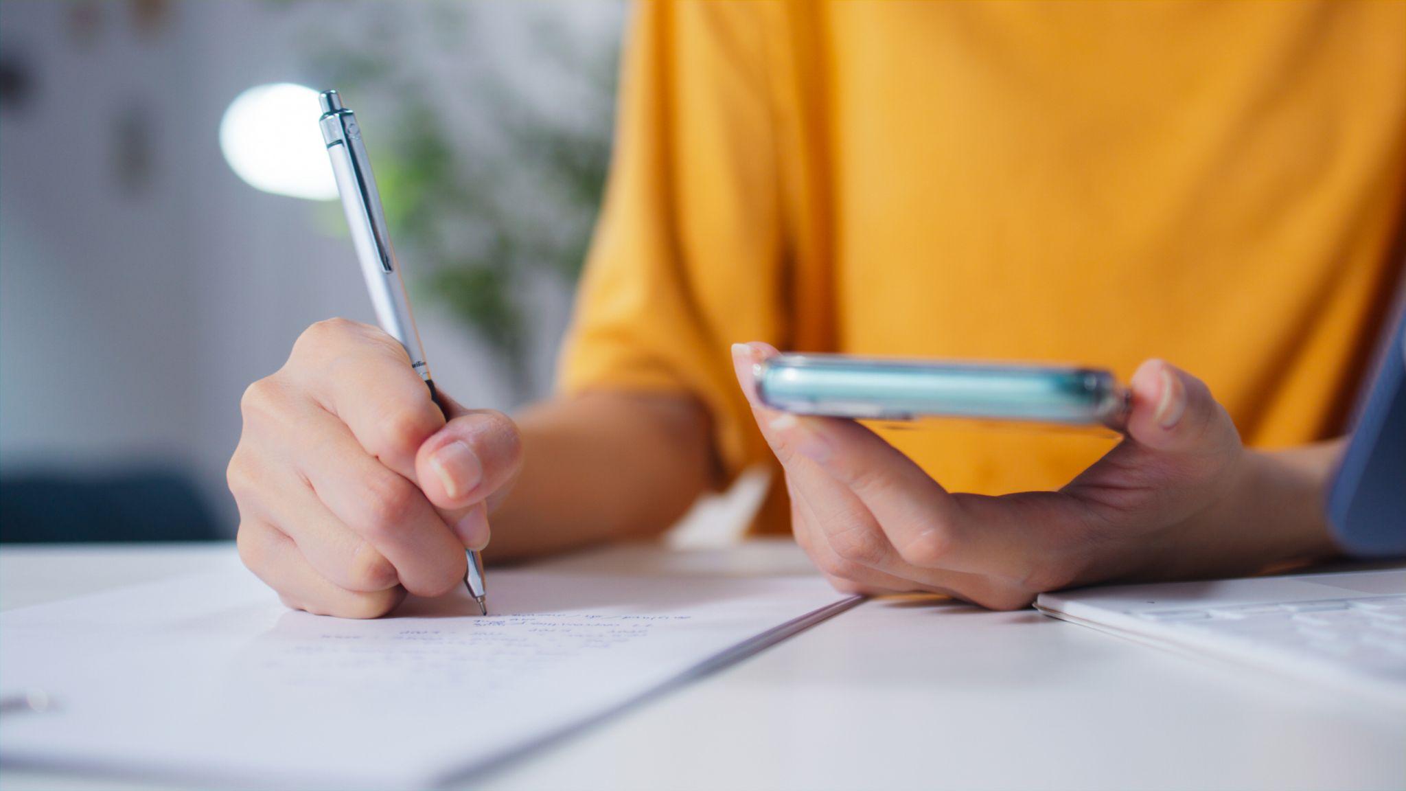 Person writing notes while holding smartphone, representing landscape maintenance budgeting or estimate process.