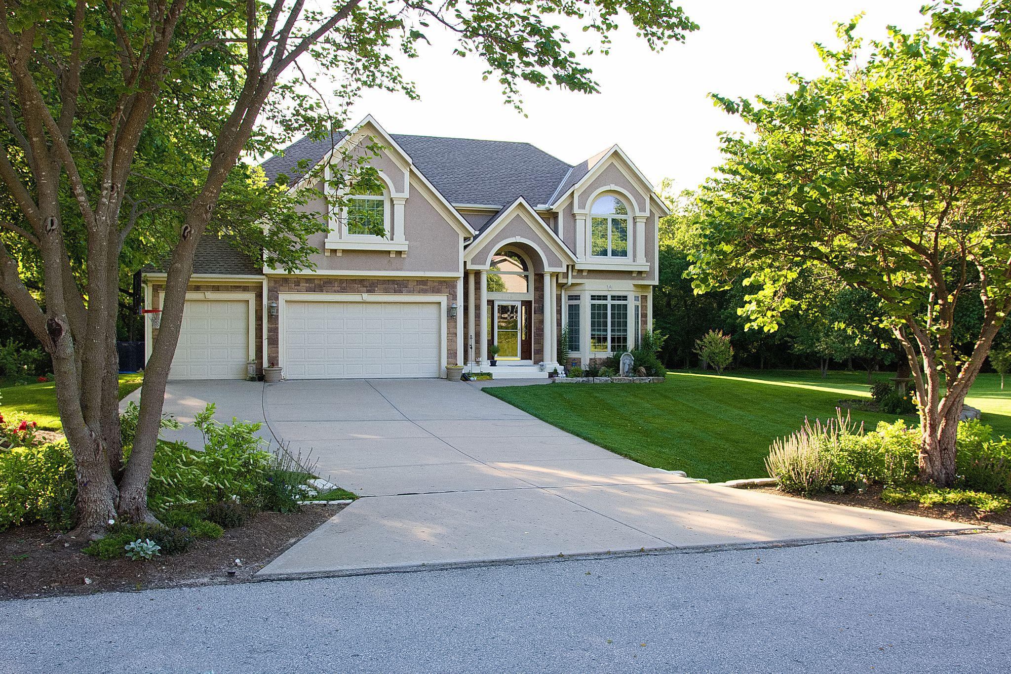 Two-story home with clean driveway, manicured lawn, and trimmed trees. Home's lawn is on a slight incline from the road. 